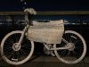 White painted ghostbike attached to a railing with flowers on the handlebar, a hand written sign in Spanish attached to it, photo taken at night, lights of NJ across the Hudson River in the background.